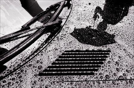 An umbrella-toting pedestrian is reflected in the rain-covered hood of his car.